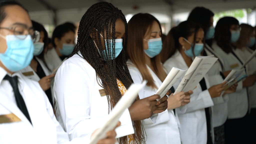 A group of students in white lab coats and masks standing and reading a ceremony program